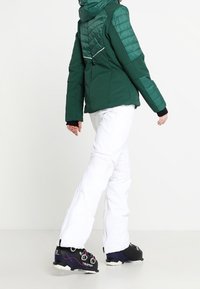 Veste de ski isolante verte avec capuche, présentant une texture matelassée, associée à un pantalon de ski blanc et des bottes de ski noires.