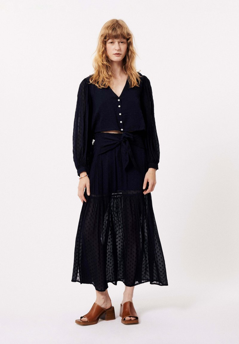 Marineblauwe twee-delige outfit met een knoopsluiting, cropped top met lange mouwen en een bijpassende lange rok met een gestructureerd stippenpatroon. Bruine hakken sandalen.