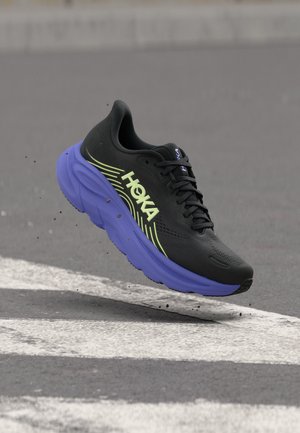 Chaussure de course noire et violette avec la marque HOKA, présentée en suspension au-dessus d'une surface pavée grise avec des lignes blanches.