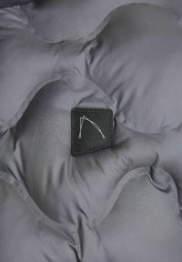 Kadar izbliza sive kviltirane tkanine s pravokutnom crnom etiketom koja sadrži minimalistički bijeli logo, s naglaskom na teksturu i detalje.