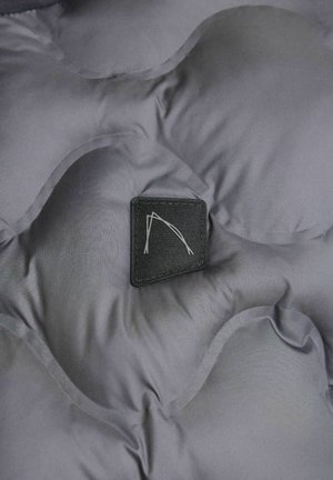 Kadar izbliza sive kviltirane tkanine s pravokutnom crnom etiketom koja sadrži minimalistički bijeli logo, s naglaskom na teksturu i detalje.