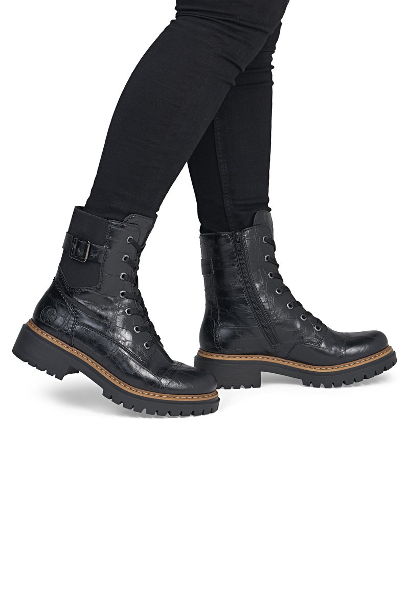 Personne portant un pantalon noir moulant et des bottes noires à lacets avec semelles épaisses et fermetures éclair sur les côtés, debout sur un fond blanc.