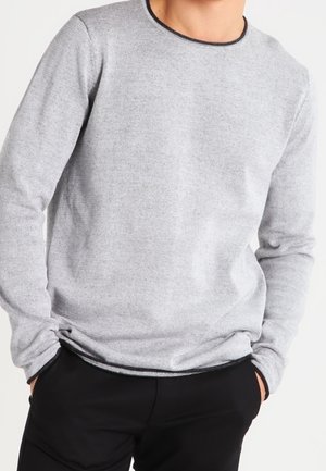 Grauer Pullover mit langen Ärmeln und Rundhalsausschnitt sowie schwarzem Saum. Weicher Stoff, taillierte Passform, getragen über schwarzen Hosen, Hände in den Taschen.