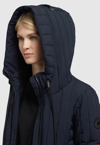 Navy puffer jacket with a large hood, quilted texture, and zip closure. Features a drawstring and a circular logo patch on the shoulder.