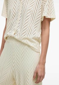 Gaiši krēmveida komplekts augšdaļai un apakšdaļai, kurā ir zigzagveida perforāciju raksts, īsās piedurknes un brīva piegriezuma forma. Gluda tekstūra un elpojošs dizains.