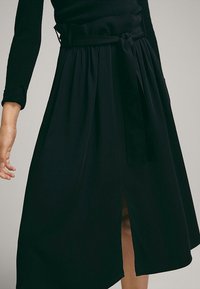 Femme portant une jupe midi noire avec une fente à l'avant et une ceinture noire assortie nouée à la taille.