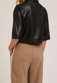Top corto in finta pelle nera con maniche corte, texture liscia e scollo pulito, abbinato a pantaloni beige su misura.