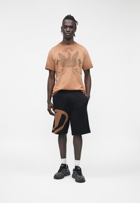 Joven con camiseta gráfica marrón, pantalones cortos negros con un diseño marrón, calcetines blancos y zapatillas negras de pie sobre un fondo blanco liso.