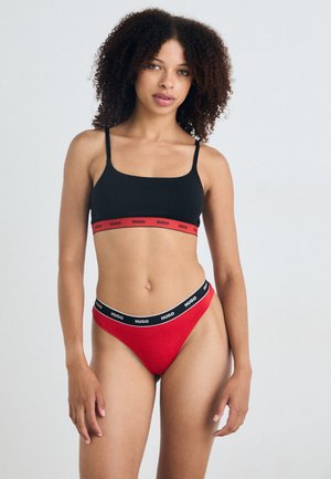 Svart bralette med rød stripe merket "HUGO," og rød truse med svart stripe merket "HUGO." Begge er laget av et glatt, tekstilmateriale.