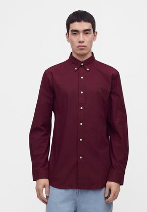 Homme portant une chemise bordeaux à manches longues boutonnée et un jean bleu clair, debout devant un fond blanc uni.