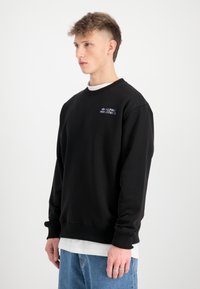 Crni dukserica s okruglim izrezom, rebrastim manžetama i donjim dijelom, s malim vezom logotipa na prsima. Nosi se preko bijele košulje, u kombinaciji s farmerkama.