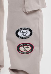 Beige broeken hebben twee patches: een ovale zwarte patch met "PEGADOR Royalty Club COLD HEARTED" en een ovale patch met een rode rand met soortgelijke tekst.