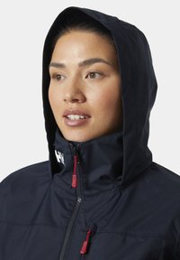 Marineblauw waterdicht jack met een hoge kraag en gevoerde capuchon. Voorzien van een voorrits met rode trekker en een opvallend logo op de kraag.