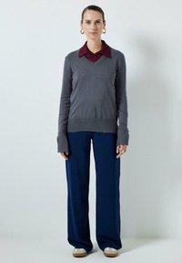 Grijze gebreide v-hals trui over een kastanjekleurig overhemd met kraag, gecombineerd met brede marineblauwe broeken en lichtgekleurde schoenen met witte accenten.