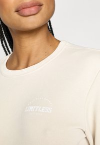 Lys beige genser med rund hals. Har en hvit rund grafisk tekst "LIMITLESS" og mykt, teksturert stoff.