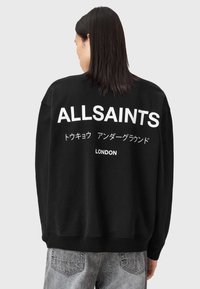 Melnā kokvilnas sporta krekls, ar lieliem baltajiem uzrakstiem "ALLSAINTS" un "LONDON" uz muguras, starp kuriem ir japāņu rakstzīmes.