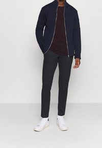 Giacca a zip navy con motivo texture, maglietta maroon a girocollo e pantaloni grigi scuri. Sneakers bianche con dettagli blu completano l'outfit.