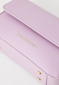 Mala lilás com um acabamento texturizado, ferragens douradas e um logótipo "Valentino" em destaque na aba frontal. Forma retangular compacta.