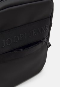 Černý nylonový batoh s texturovaným popruhem a vdechovaným logem "JOOP! JEANS" a zipovou přední kapsou se hladkým taháčkem.