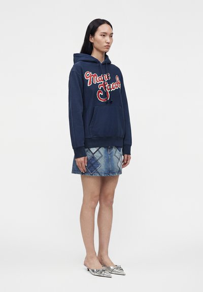 Γυναίκα με ναυτικό μπλε φούτερ Marc Jacobs, τζιν patchwork μίνι φούστα και ασημένιες μυτερές μπαλαρίνες, σε λευκό φόντο.