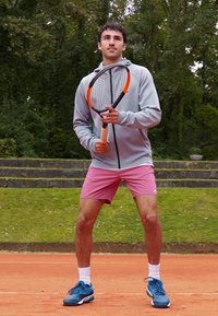Idrottare i en ljusgrå zip-up jacka och rosa shorts, som håller en tennisklova med orange detalj, stående på en grusplan.