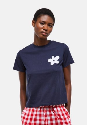 Femme aux cheveux courts portant un t-shirt bleu marine avec un motif de fleur blanche et un pantalon à carreaux rouges et blancs sur un fond uni.