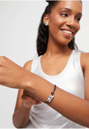 Mujer sonriente con una camiseta sin mangas blanca que lleva una pulsera de gema azul con un cordón marrón ajustable en su muñeca izquierda.