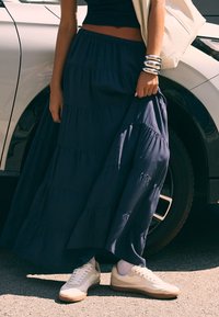 Žena stojí pri bielom aute, oblečená v námornícky modrej maxi sukni s volánmi, čiernom crop top, bielych teniskách, bielych ponožkách a strieborných náramkoch.