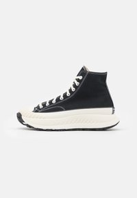 Bottine noire avec lacets blancs, tige en toile, bout arrondi avec protection en caoutchouc, semelle blanche épaisse et semelle extérieure noire texturée.
