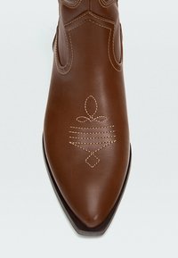 Brun läderstövel med spetsig tå och dekorativ beige söm i abstrakta geometriska mönster på den övre framsidan.