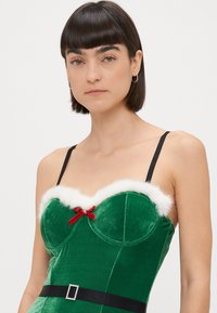 Robe en velours vert avec un corsage ajusté, des bordures en fourrure blanche, un accent en nœud rouge et des bretelles noires. Dotée d'une ceinture décorative noire avec une boucle.