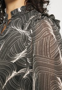 Blusa de patrón en blanco y negro con tela fluida, que presenta un cuello alto con volantes y una textura semi-translúcida. Los detalles incluyen delgadas líneas blancas.