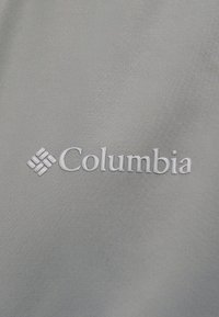 Λογότυπο της μάρκας Columbia με έμβλημα σε σχήμα διαμαντιού κεντημένο με λευκό νήμα σε λείο γκρι ύφασμα.