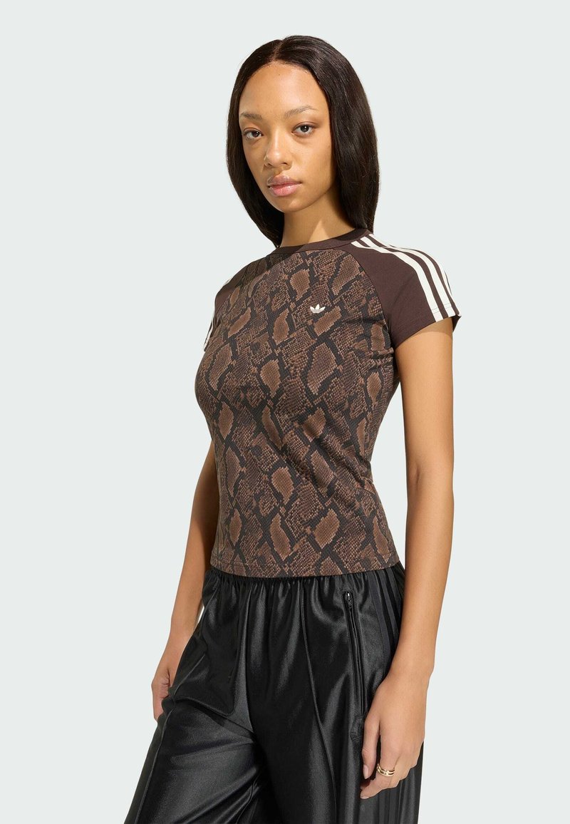 Femme portant un haut marron à manches courtes avec un imprimé serpent et des rayures blanches sur les manches, ainsi qu’un pantalon noir ample, debout devant un fond uni.