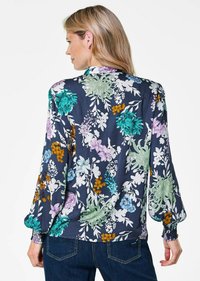 Blusa floreale con maniche lunghe in tessuto blu scuro; presenta grandi fiori in teal, viola e arancione; polsini e orlo elasticizzati.