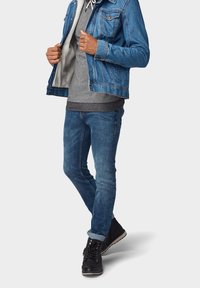 Denim jack in middelblauw, gecombineerd met een grijze hoodie eronder. Passende blauwe jeans en zwarte sneakers benadrukken de casual outfit.