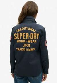 Navy blue work jacket with golden embroidered text on the back, featuring accents in red and white on the sleeves.