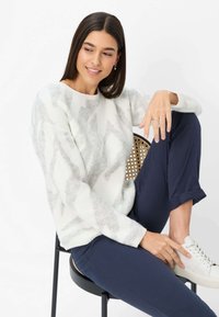 Donna con capelli scuri indossa un maglione bianco con motivo, pantaloni blu navy, scarpe da ginnastica bianche, seduta su una sedia con una gamba sollevata, sorridente di lato.