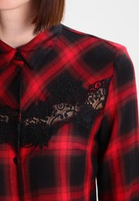 Chemise à carreaux rouge et noir avec un col; dotée de détails en dentelle avec des motifs floraux sur la poitrine et des fermetures par boutons.