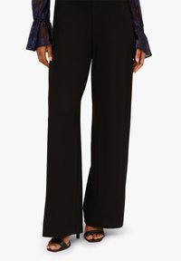 Czarne spodnie o szerokich nogawkach z gładką fakturą, z rozcięciem z przodu, w połączeniu z prostymi czarnymi sandałami. Bluzka ma przezroczyste, niebieskie akcenty.