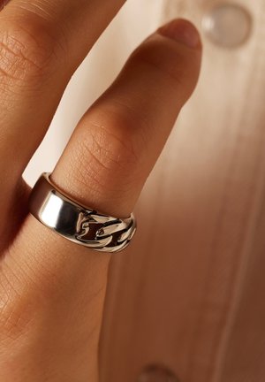 Silberner Ring mit einem polierten Band und einem verflochtenen Kettendesign. Glatte Textur mit glänzendem Finish, am Fingerspitze getragen.