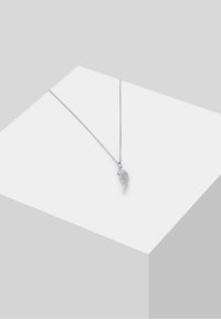 Collier en argent avec un pendentif en forme d'aile d'ange détaillée, posé sur une surface d'exposition blanche inclinée.