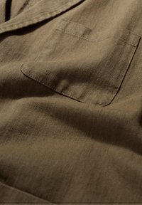 Țesătură de culoare maro cu model în zigzag, având o textură netedă, care include un buzunar frontal și cute subtile.