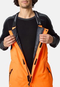 Oranje waterdichte overalls met verstelbare zwarte banden, een ritssluiting aan de voorkant en een zwarte binnenvoering. Voorzien van zijzakken en een stevig ontwerp.