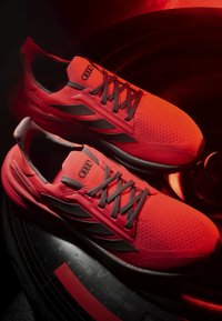 Paire de chaussures de course Adidas Ultraboost rouges avec lacets noirs et logo Audi, placée sur un pneu noir sous un éclairage rouge.