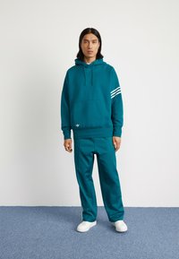 Felpa teal con cappuccio e tasca a marsupio, con strisce bianche sulla manica sinistra. Abbinata a pantaloni teal coordinati e sneakers bianche.
