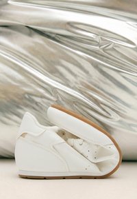 Bílý tenisek přehnutý na polovinu, aby ukázal svou flexibilní podrážku s gumovou barvou na spodní části, na pozadí lesklé stříbrné látky.