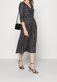 Robe noire étincelante avec un décolleté en V et des manches longues, taille cintrée et longueur midi. Portée avec des talons noirs à brides et un petit sac à main.