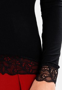 Sort ribbet langærmet top med blomsterlacemønster på manchetterne og kanten, parret med røde bukser.