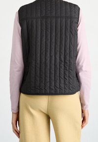 Chaleco negro acolchado con patrón de acolchado vertical, escote redondo y sin mangas, usado sobre una camiseta de manga larga rosa y pantalones beige.